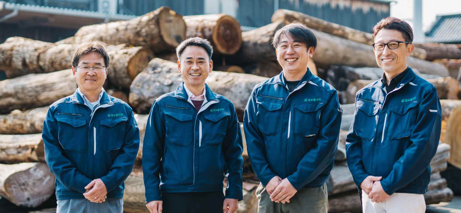 積み上げられた原木の前で、木材アドバイザーの男性4人が笑っている写真。